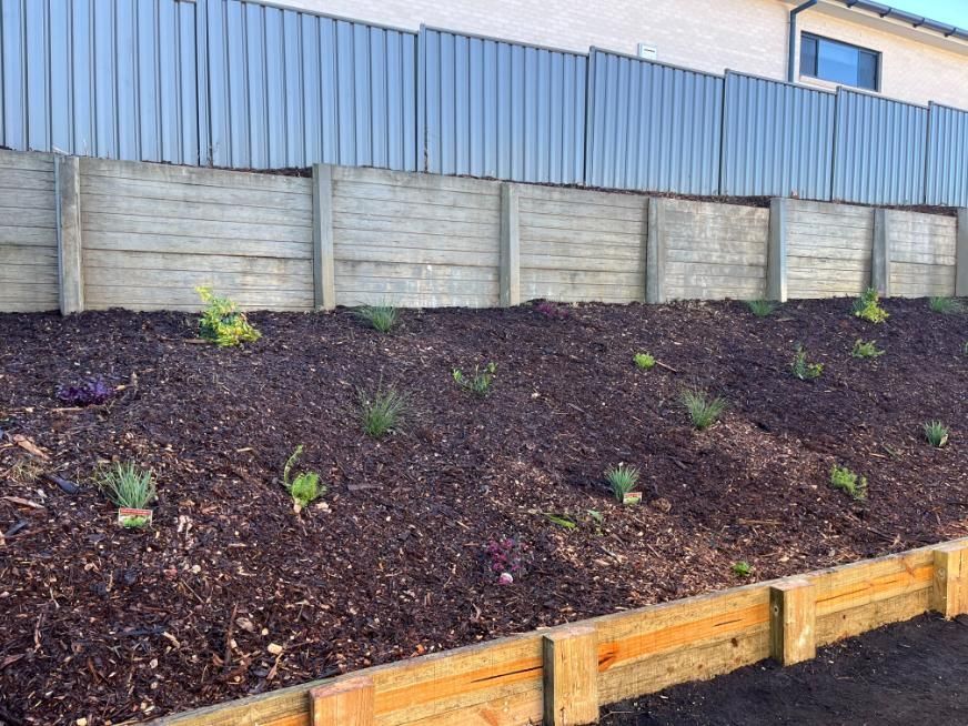 A Concrete Wall With A Wooden Retaining Wall And A Pile Of Mulch In Front Of A House — Lewis Landscapes In Wauchope, NSW