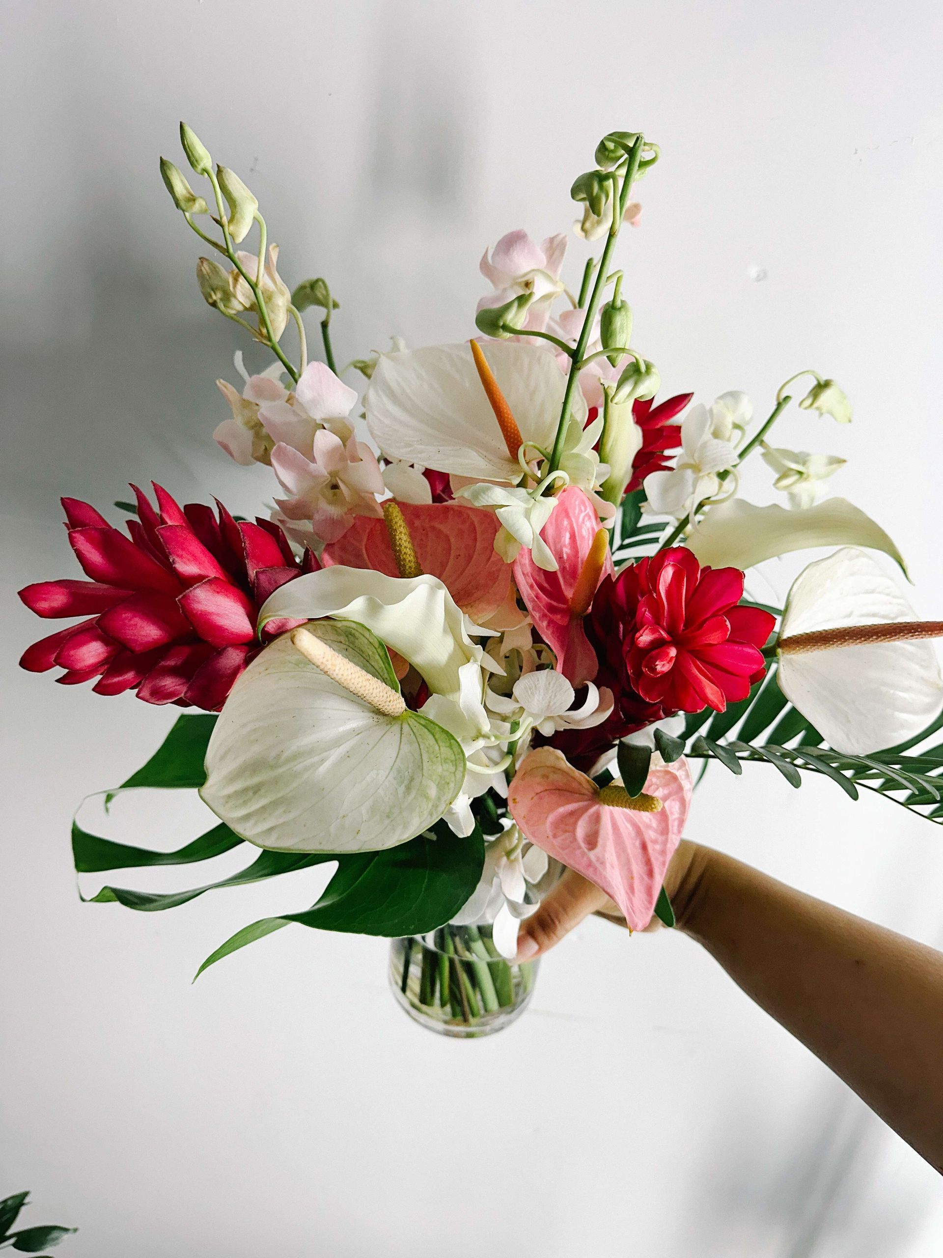 Vibrant bouquet of red, pink, and white tropical flowers in a clear glass vase, held by a person.