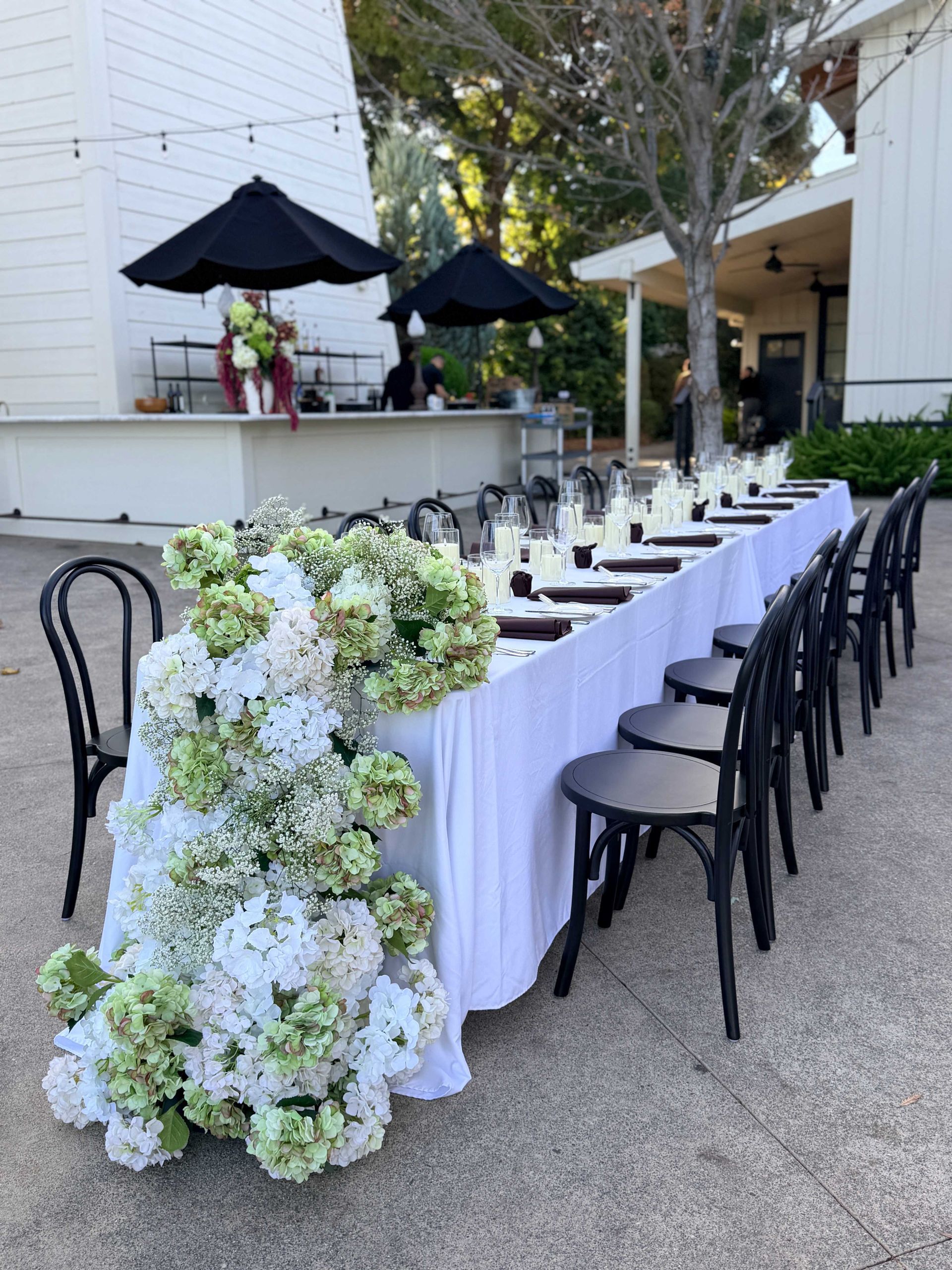 Outdoor table setting with white tablecloth, black chairs, and floral arrangements.
