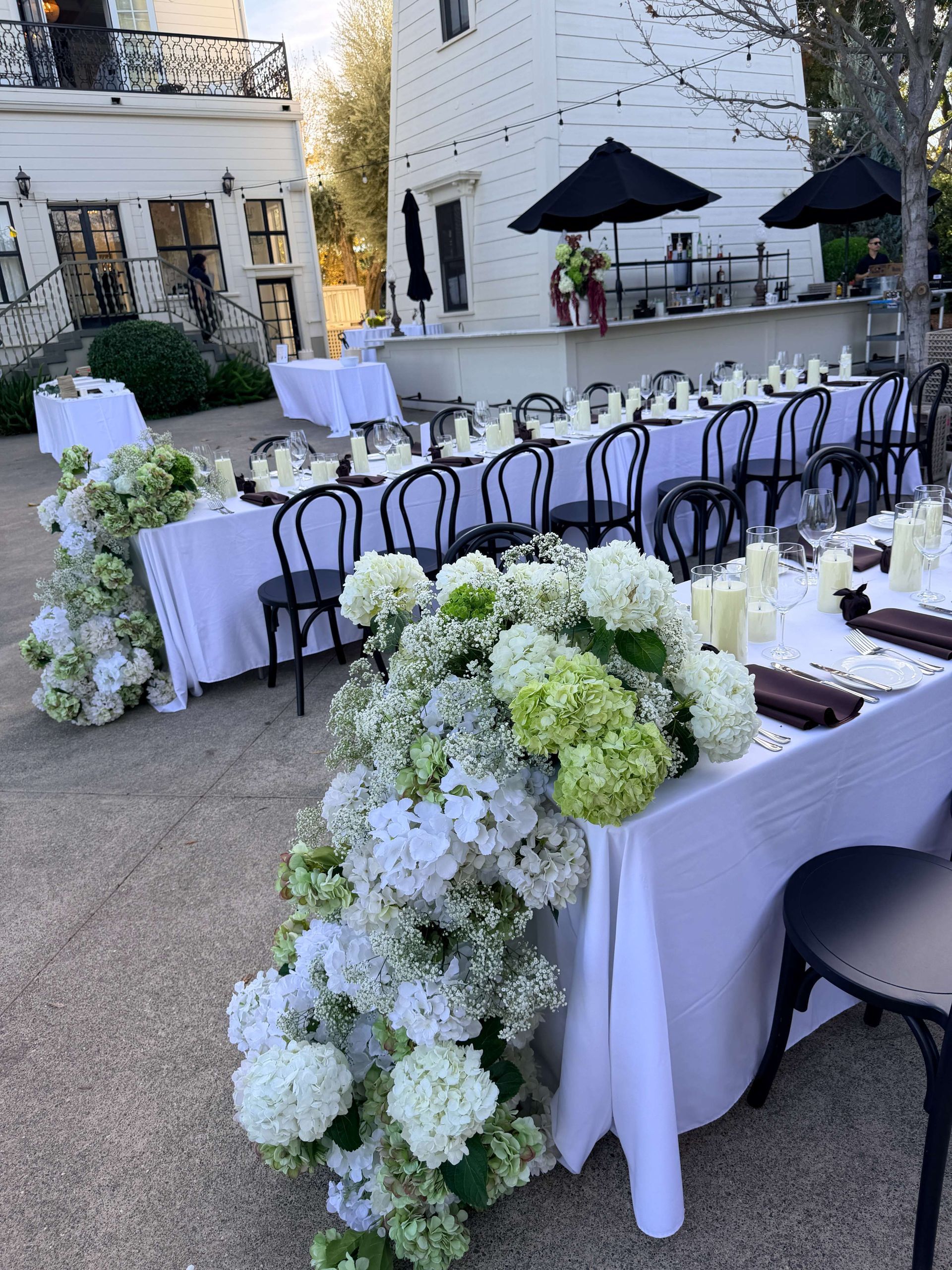 Outdoor reception tables decorated with white flowers, white tablecloths, and black chairs.