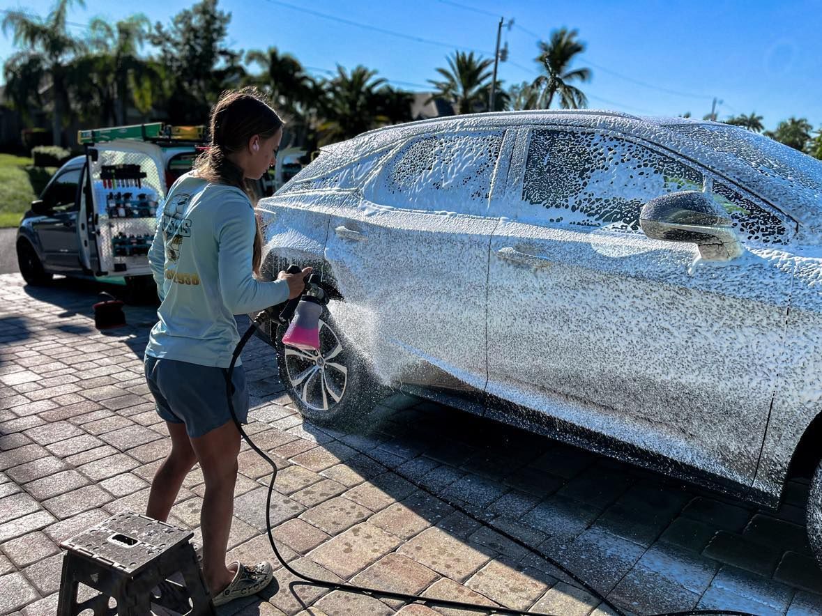 A girl is washing a red sports car in a parking lot.