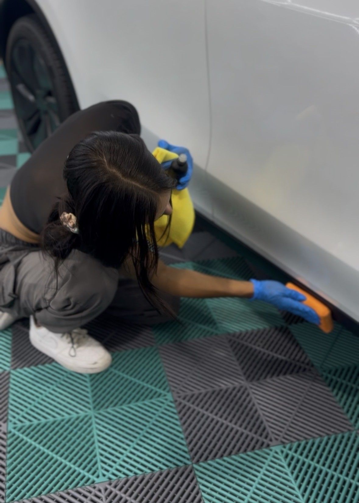 Person crouches beside a white car, wiping the lower panel with an orange cloth on a patterned floor.