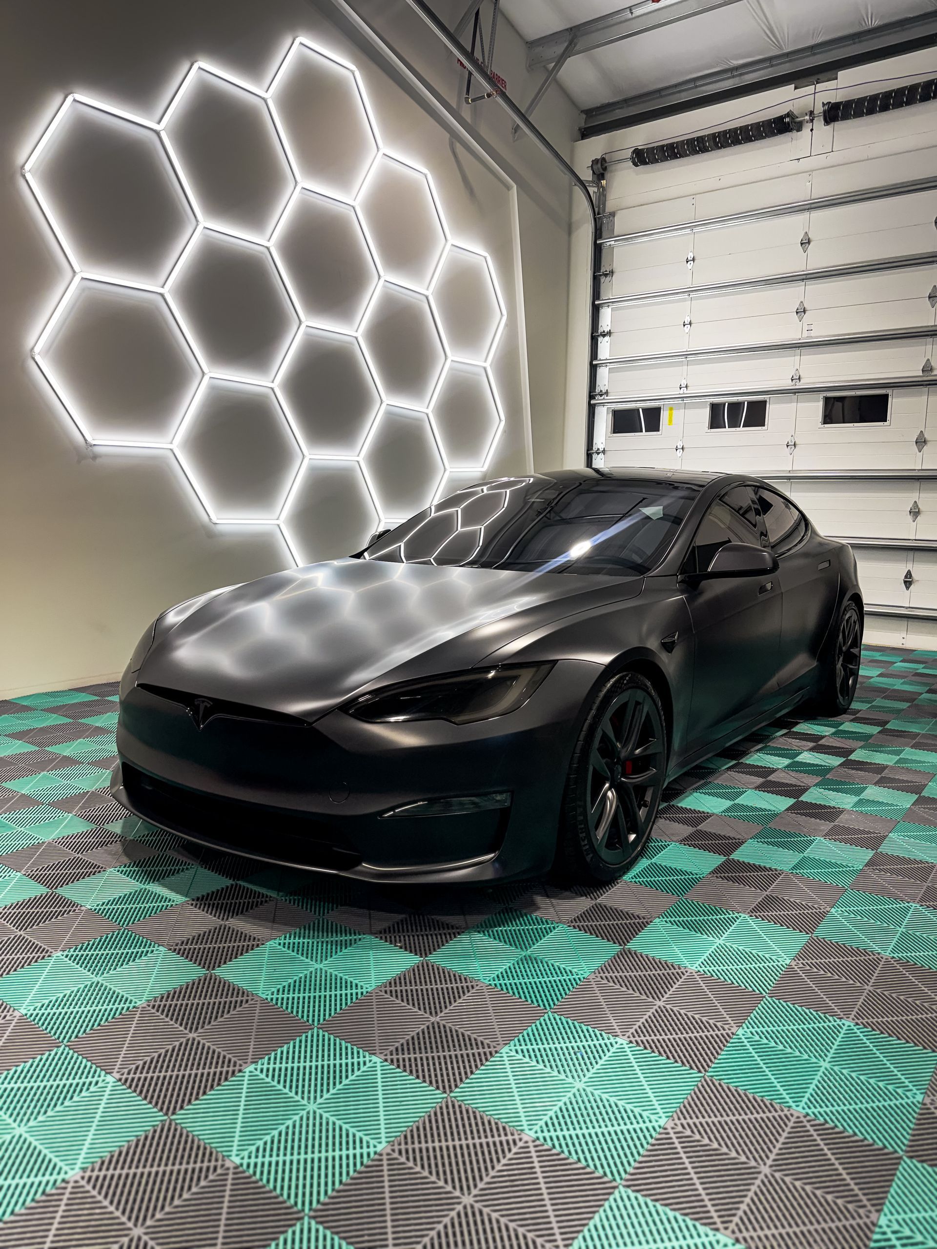 Black sports car in a garage with bright hexagon wall lights and patterned floor.