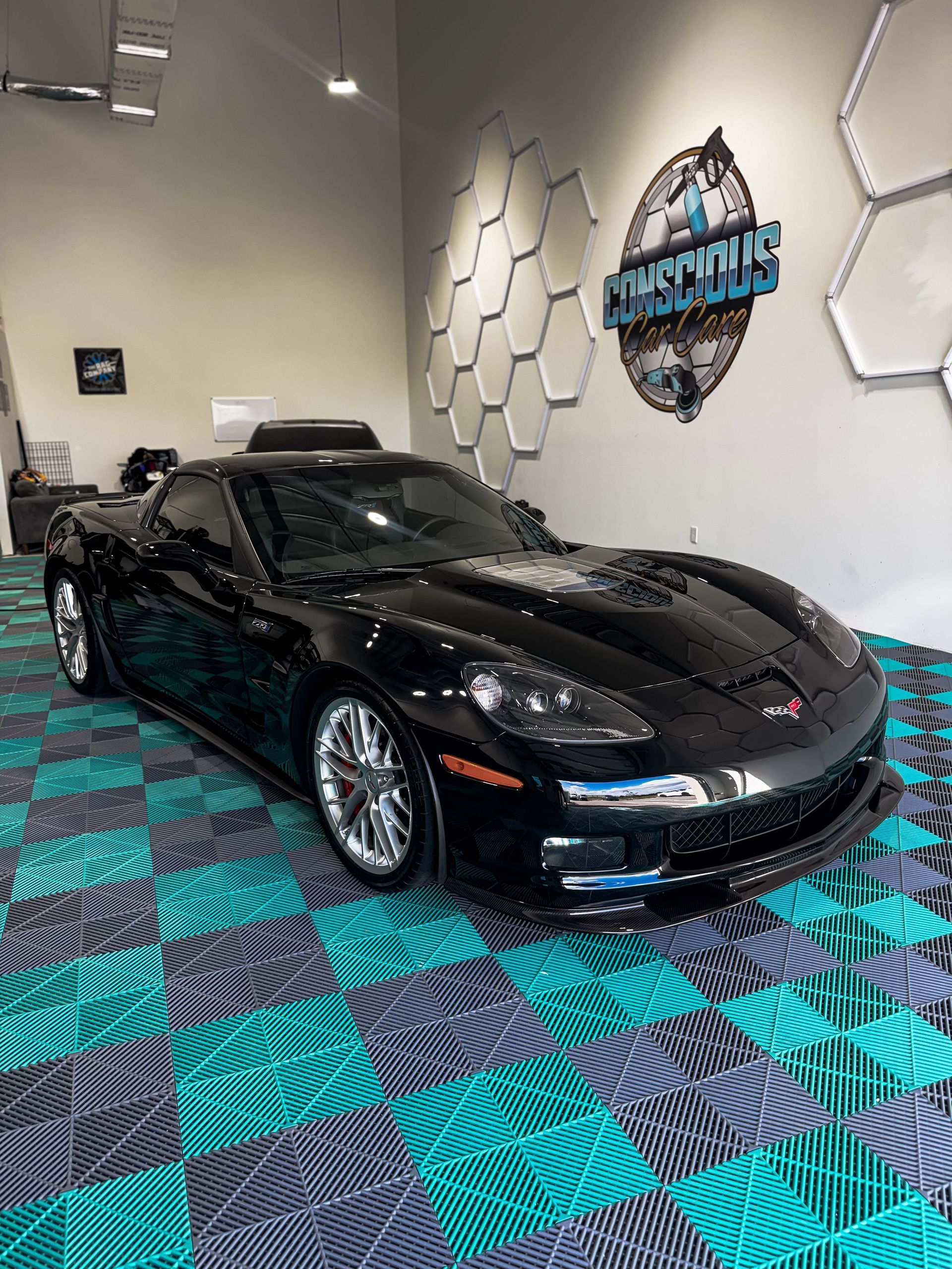 Black sports car displayed in a showroom with teal-and-black geometric carpet and hexagon wall decor
