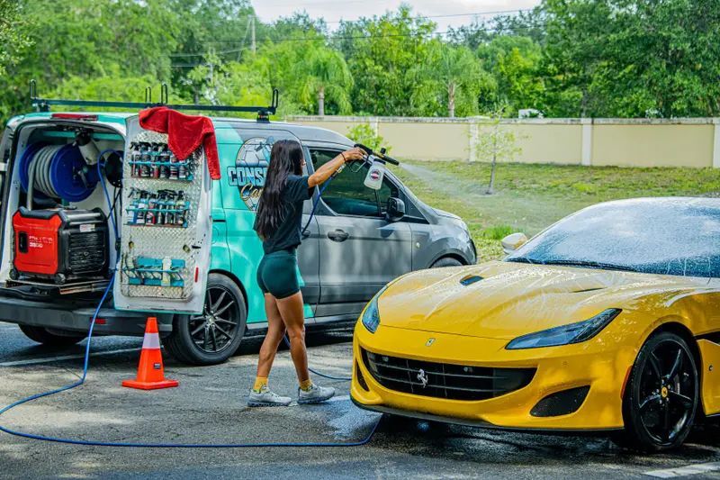 Person rinses a gray car beside a yellow sports car at a car wash.