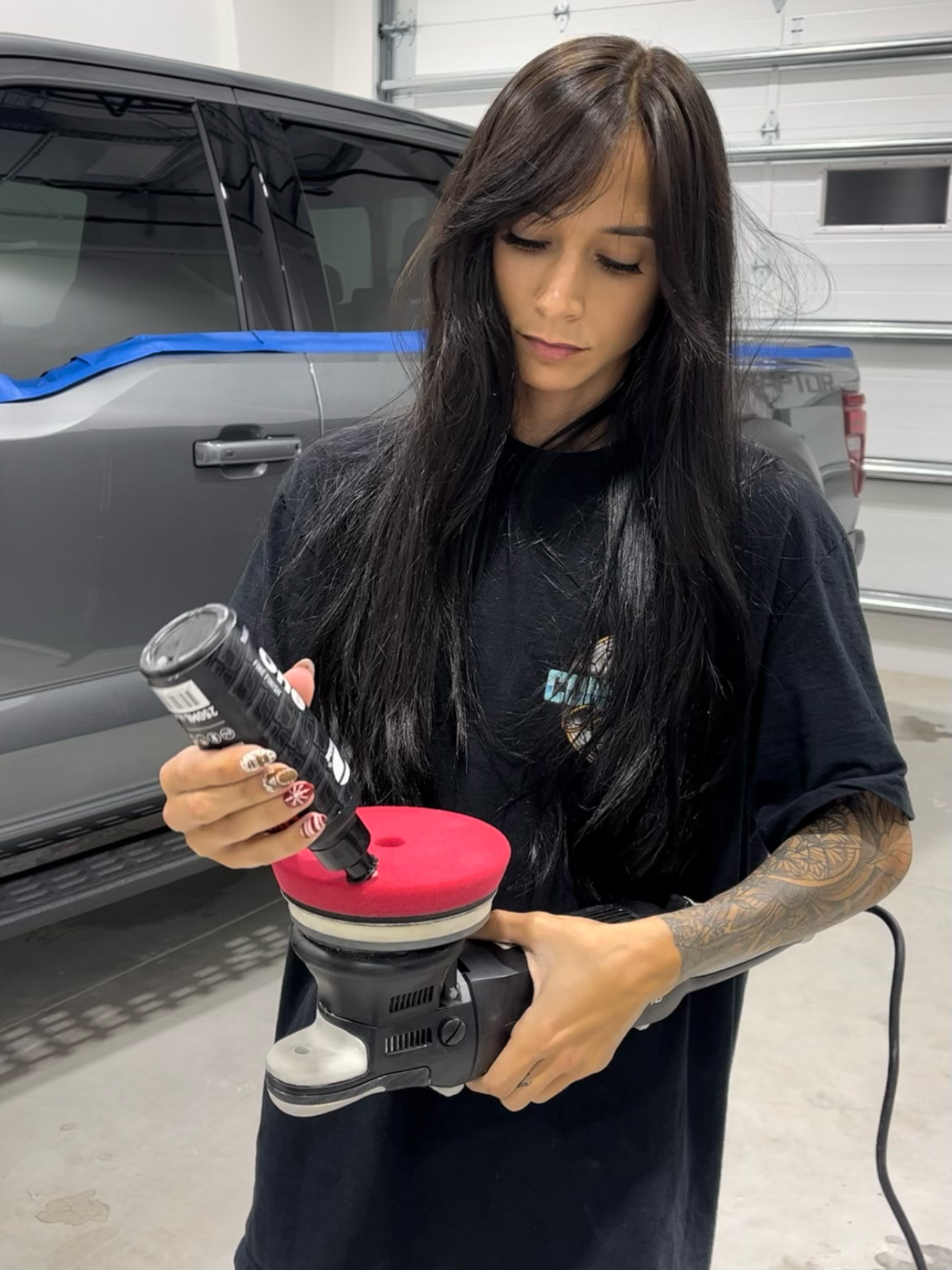 Woman polishing car panel with power buffer in a garage, wearing a black shirt.