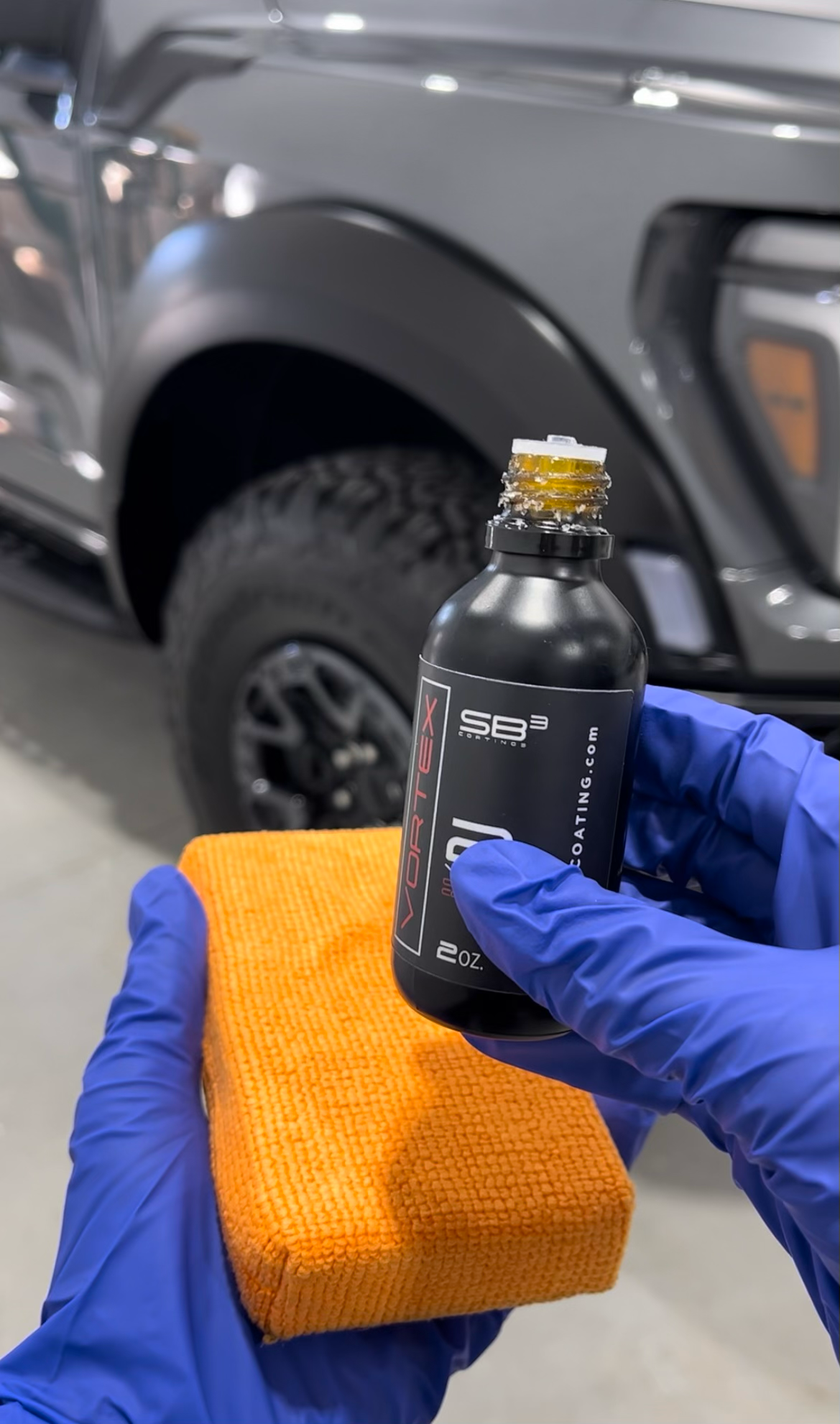 Hands in blue gloves holding ceramic coating bottle and applicator pad, next to car tire.