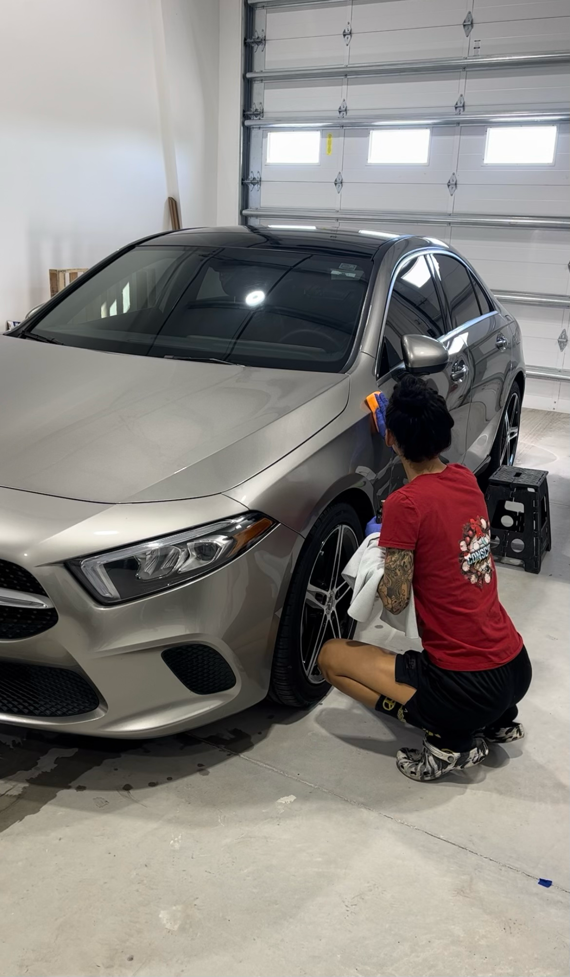 Person cleaning the front wheel of a silver Mercedes-Benz sedan inside a garage.