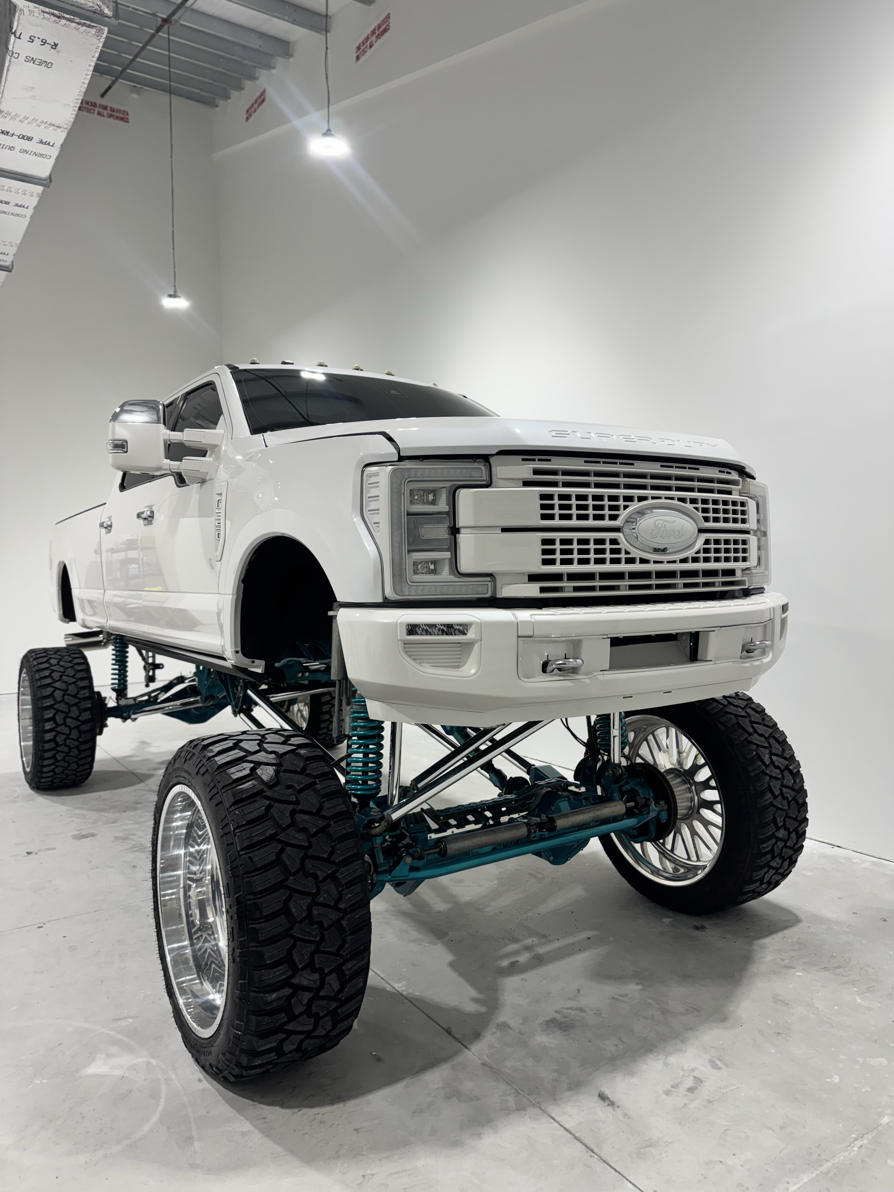 White lifted Ford truck with large chrome wheels and teal suspension, in a white studio.