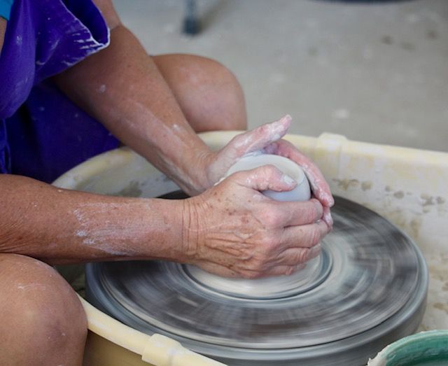 A potter's hands shaping a clay pot on a spinning wheel; close-up, hands covered in clay.