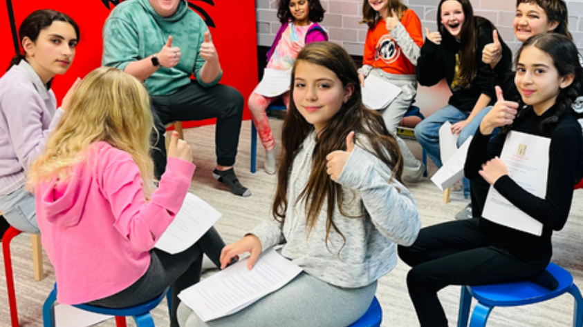 Group of children giving thumbs up, seated with papers on blue chairs in a room with red background.