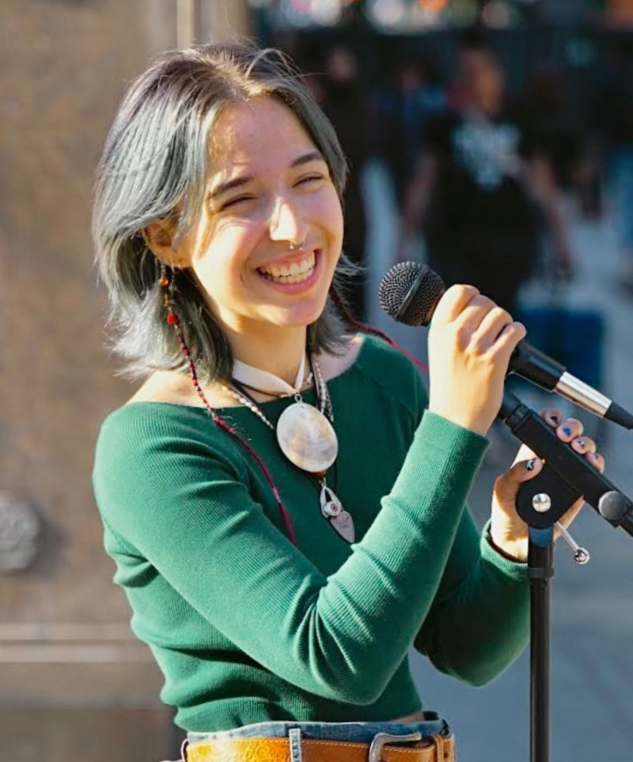 Woman with blue-grey hair, smiling, singing into a microphone. She wears a green top and necklace, performing outdoors.