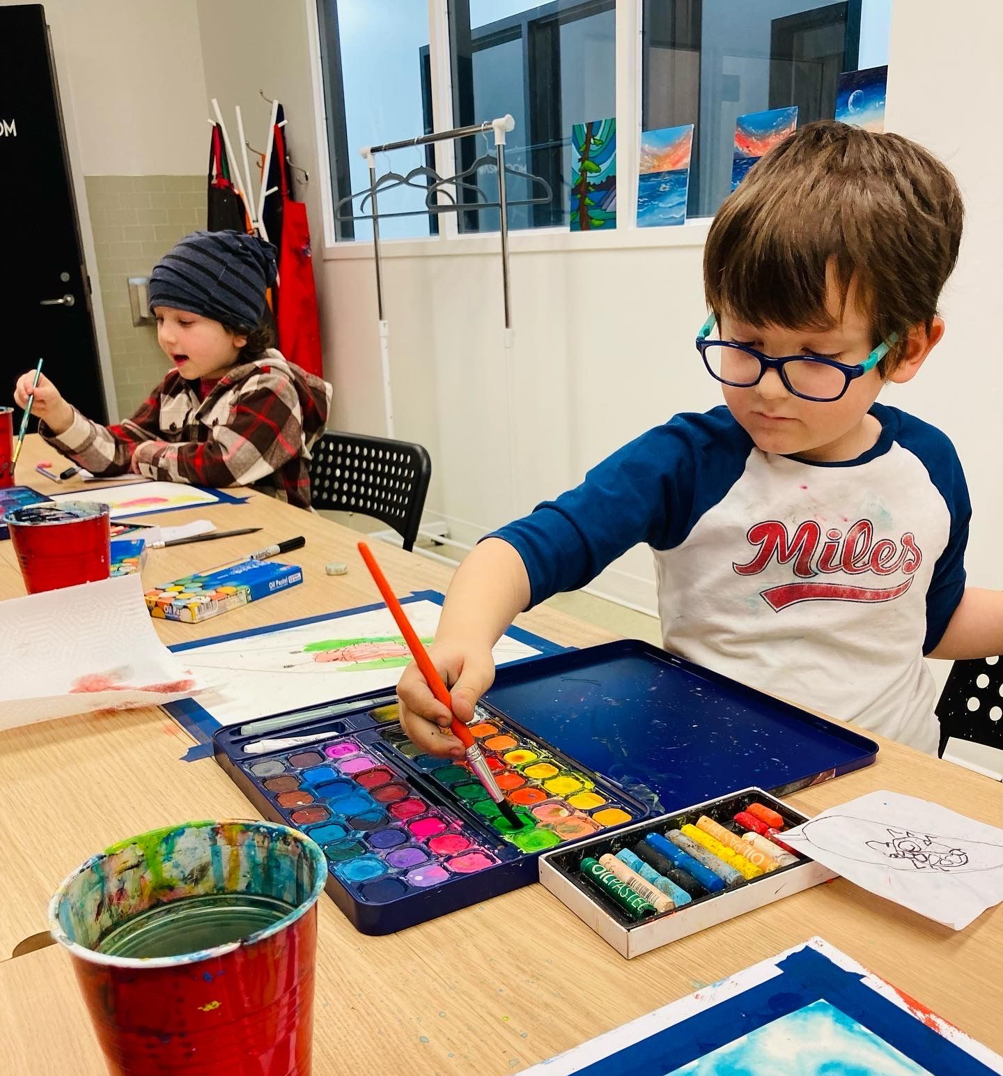 Two children painting at a table in a classroom. One uses watercolors, the other, crayons. Red cup, color palettes visible.