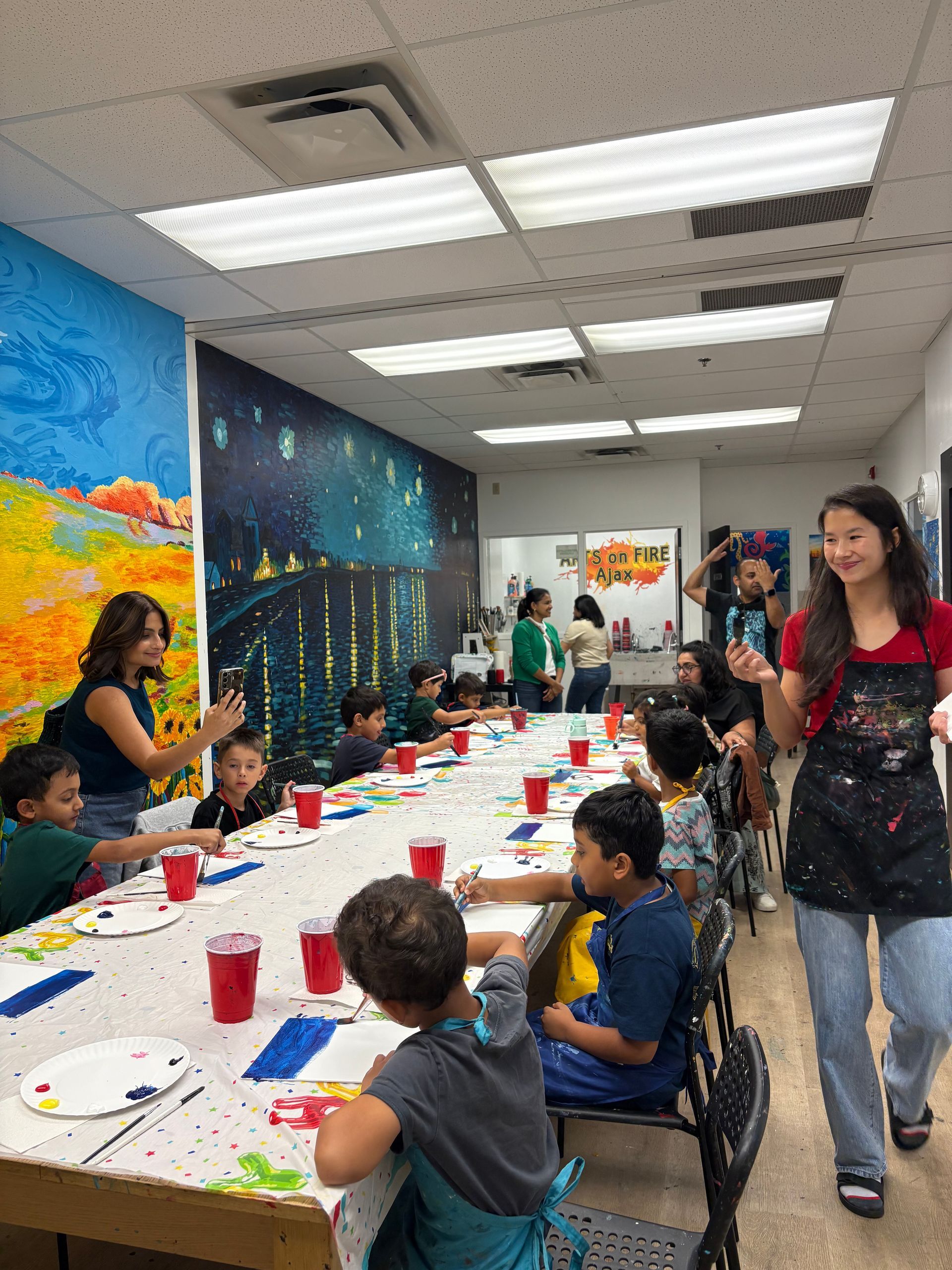 Children painting at a long table in an art class. A mural decorates the wall.