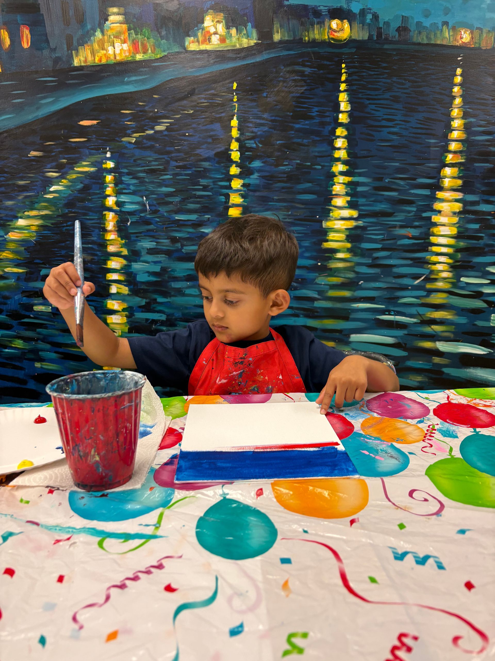 Young child painting, seated at a craft table. Artwork features blue, white, and red bands. Background is a night cityscape.