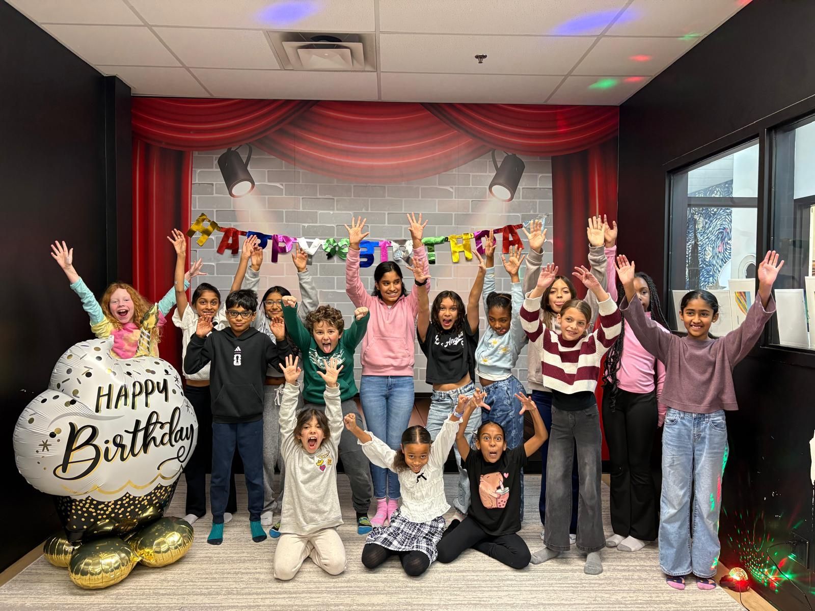 Group of children celebrating birthday, arms raised in a room with a theater backdrop and balloons.