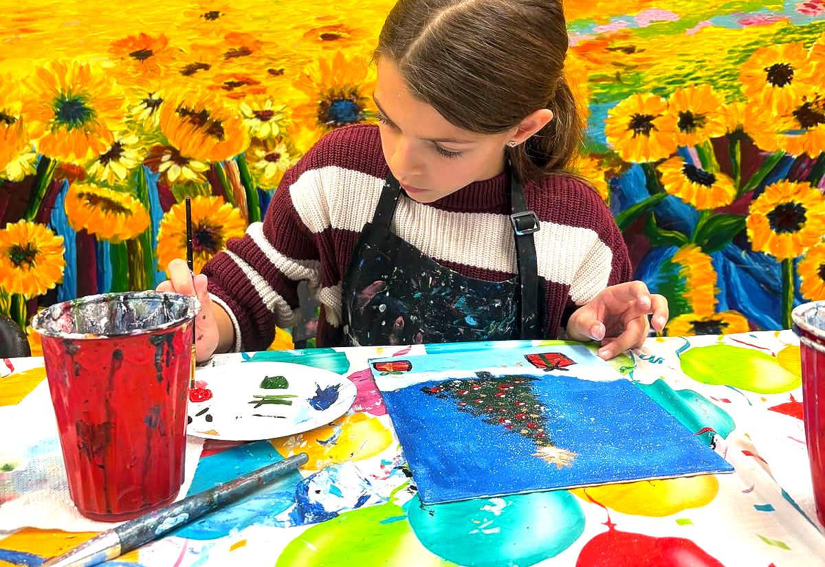 Girl painting at a table with sunflowers in the background, holding a paintbrush and wearing an apron.