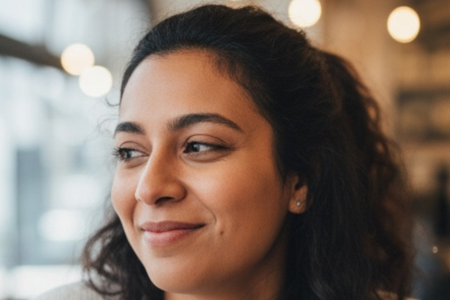 A person with dark, wavy hair looking off to the side with a gentle, thoughtful smile against a blurred, warm background.