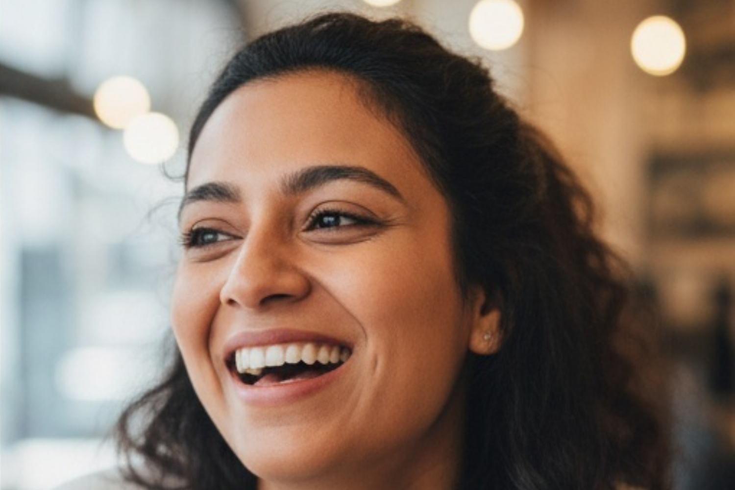 Close-up of a person with curly dark hair smiling broadly with a cheerful expression in a softly lit, indoor setting.