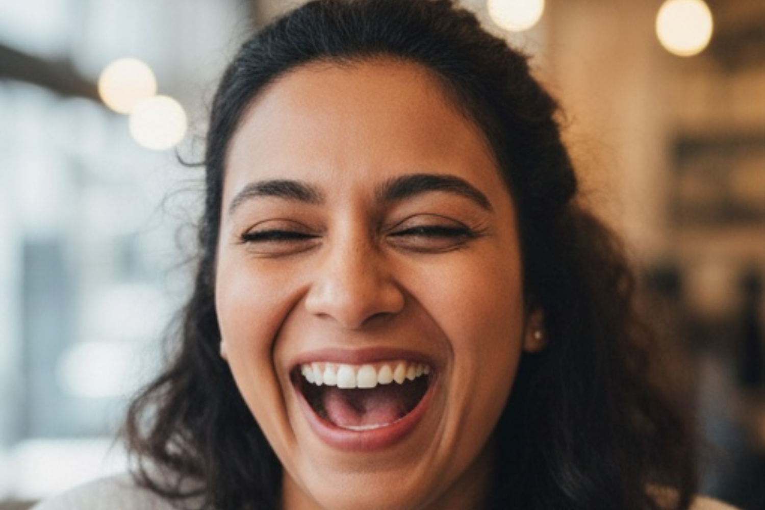 A person with dark, wavy hair laughing joyfully with eyes closed, set against a blurred, warmly lit background.
