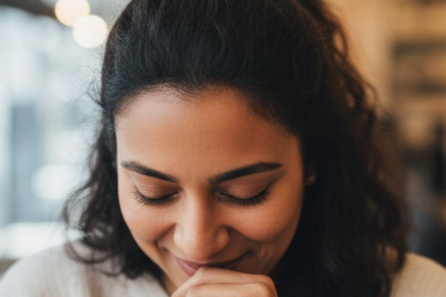 A person with curly hair looks down with a gentle smile, their hand resting near their mouth in a soft, candid moment.