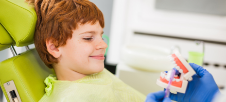 A young boy is sitting in a dental chair while a dentist holds a model of his teeth to demonstrate proper brushing technique.