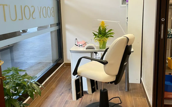 Postazione per manicure da salone con sedia e scrivania bianche, piante e porta in vetro con logo.