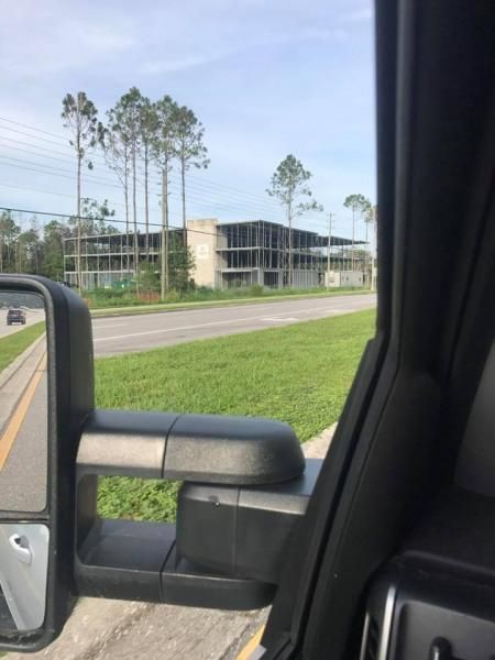 View from a car window of a construction site with a partially built concrete building and tall trees beside a road