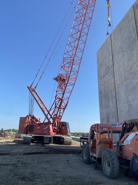 Red lattice-boom crane beside a concrete wall at a construction site, with a utility vehicle in front