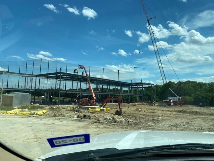 Construction site with steel frame building, crane, and dirt lot under blue sky, viewed from a car windshield