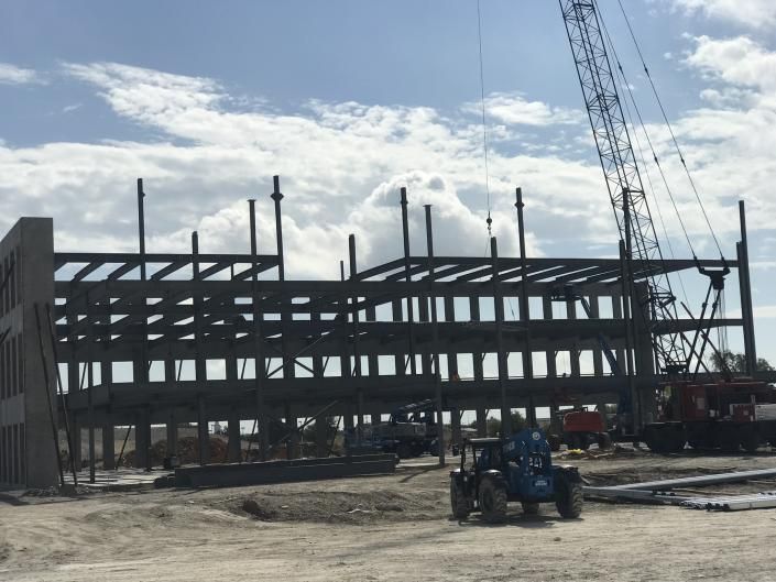 Steel-frame building under construction with a crane and blue equipment in front under a cloudy sky