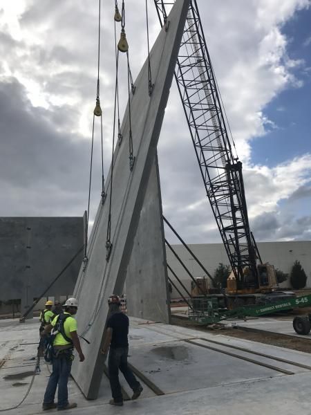 Workers guiding a large concrete wall panel beside a crane at a construction site under cloudy skies
