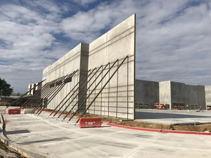 Concrete sound barrier wall under construction beside a road, with metal supports and red barriers under a cloudy sky