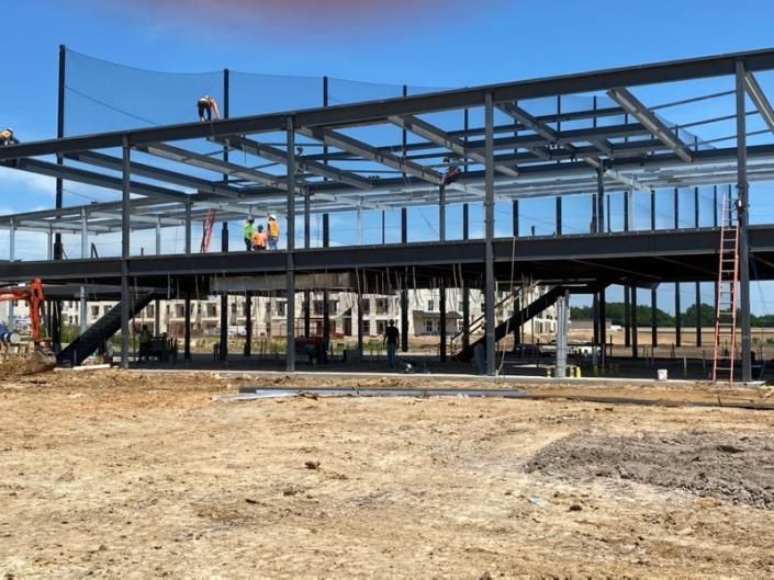 Steel building frame under construction on a dirt lot, with workers on the structure and a ladder at right.