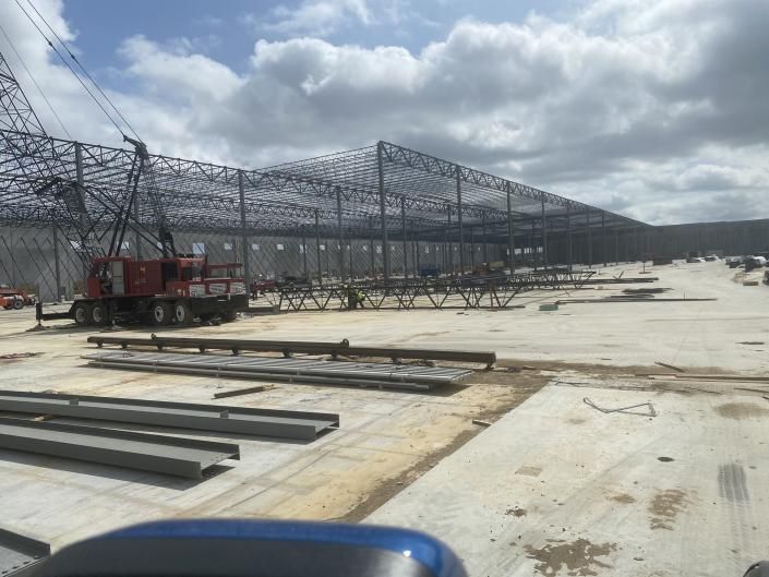 Steel truss structure under construction on a concrete site, with a red truck and cloudy sky.