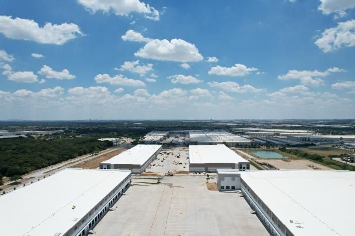 Aerial view of large white warehouse roofs in an industrial complex under a blue sky with scattered clouds