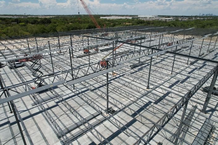 Steel roof framing under construction on a large building site with workers and scaffolding visible.