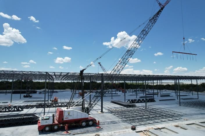 Crane lifting steel beams at a construction site beside water under a blue sky