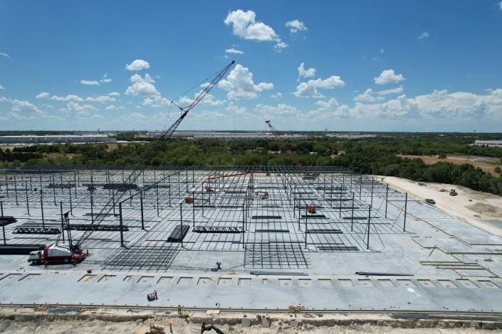 Construction site with steel foundation grid, cranes, and blue sky over a flat landscape