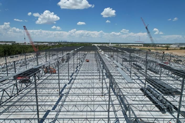 Large steel framework under construction on a wide site beneath a bright blue sky with clouds