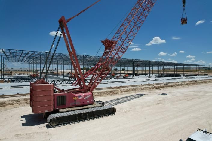 Red lattice boom crane at a construction site beside a large steel building frame under a blue sky.