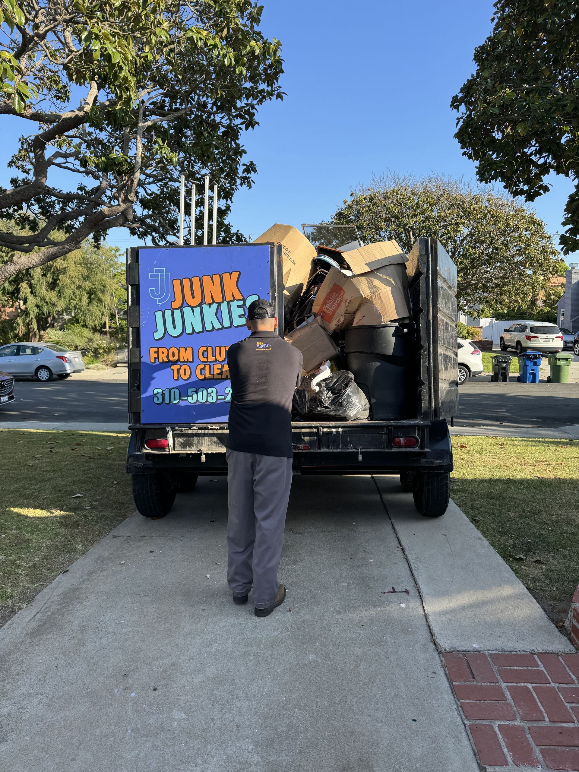 Person loading boxes into a truck with a colorful “JUNK” sign on the back, parked on a suburban street.