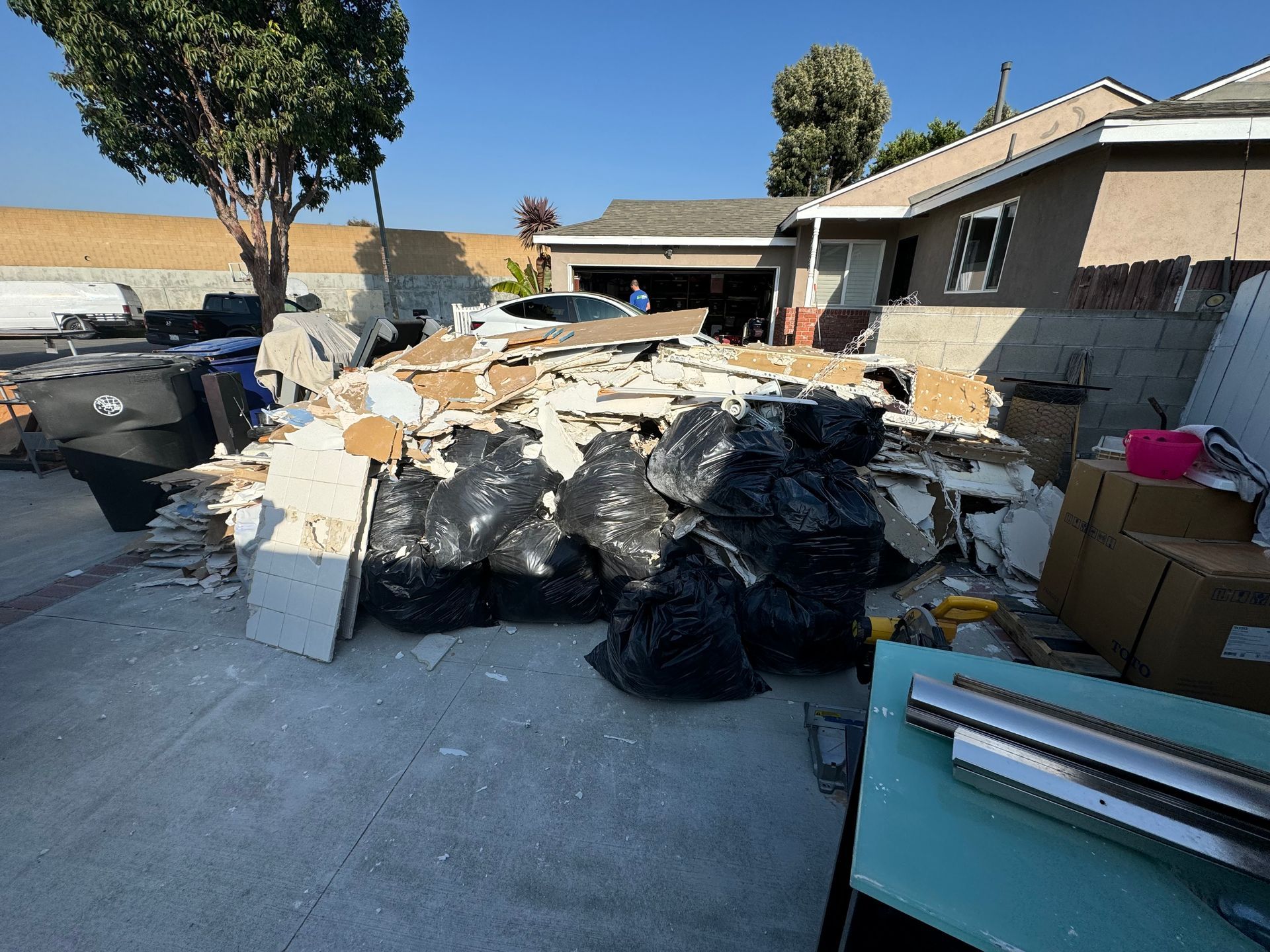Large pile of trash and debris piled in a driveway beside a house, with trash bags and bins nearby.