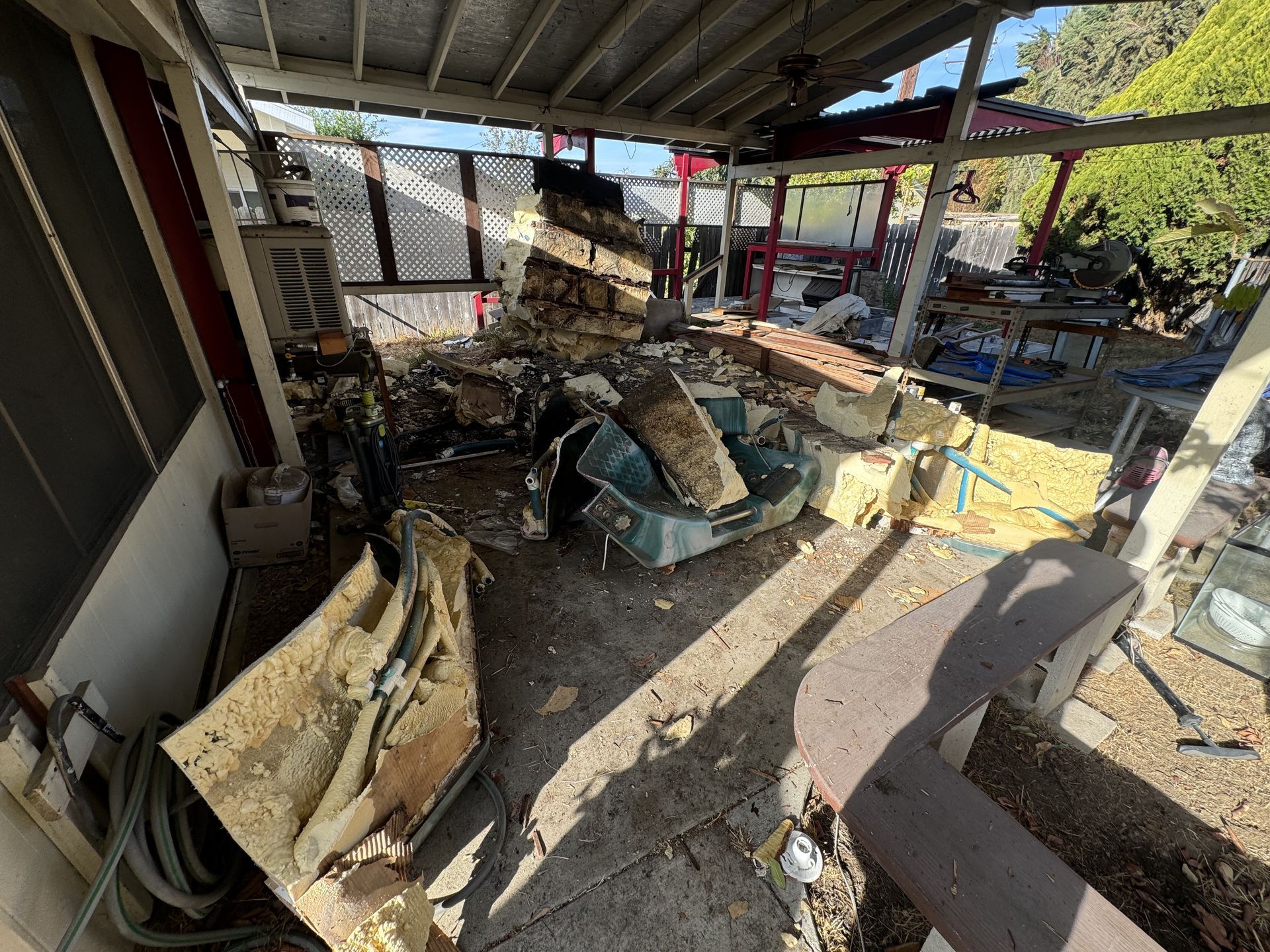 Damaged outdoor patio with broken furniture and a jacuzzi, debris, and scattered wood under a covered roof.