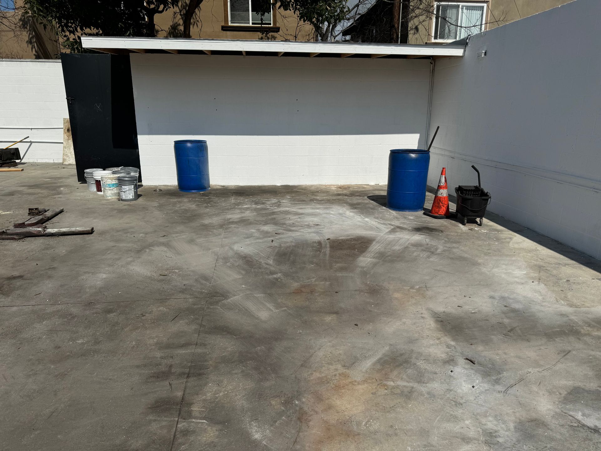 Empty concrete parking area with white walls, two blue barrels, and an orange cone by the right wall broom swept.