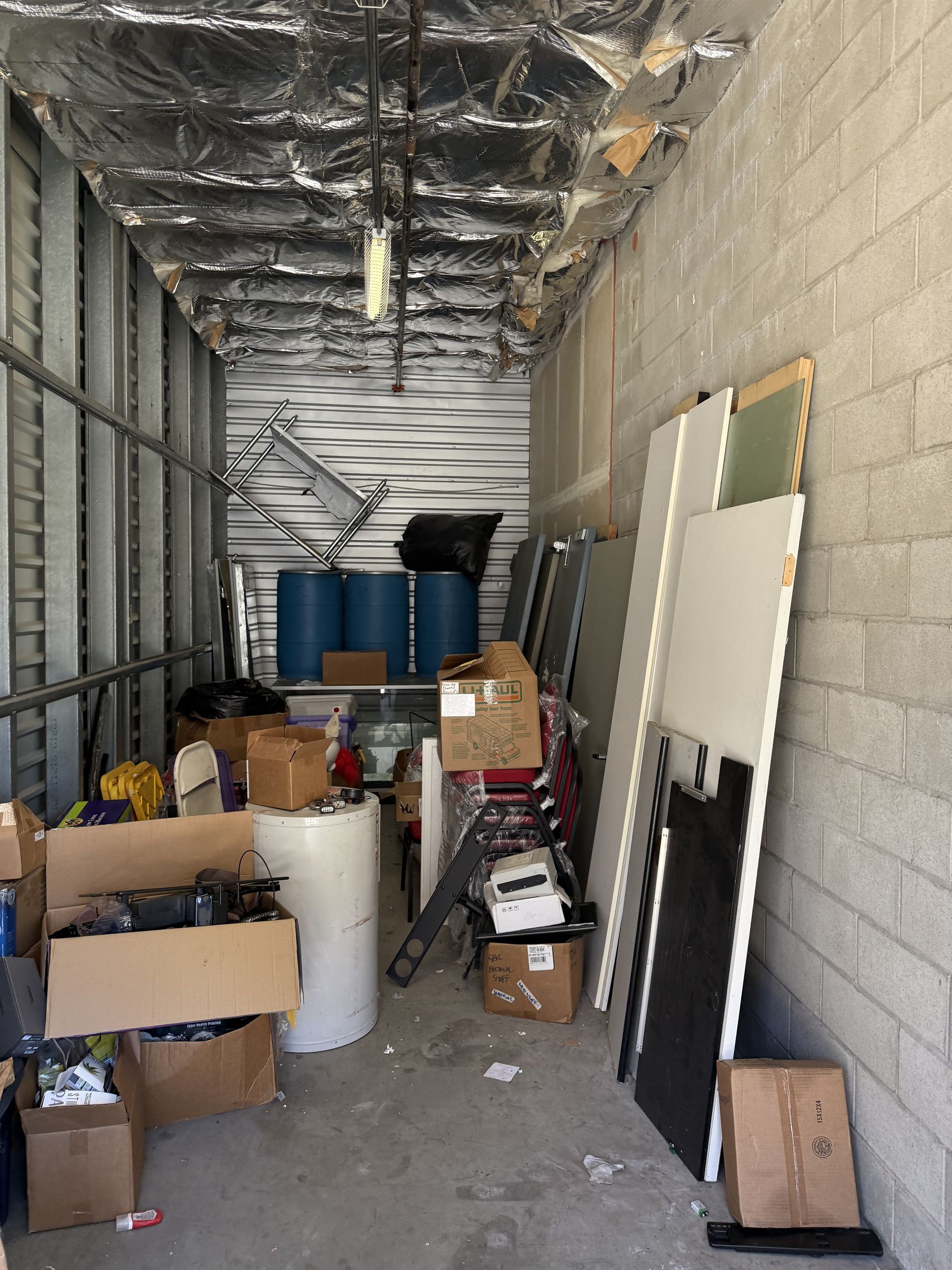 Cluttered storage corridor with boxes, barrels, and leaning panels under a metal ceiling