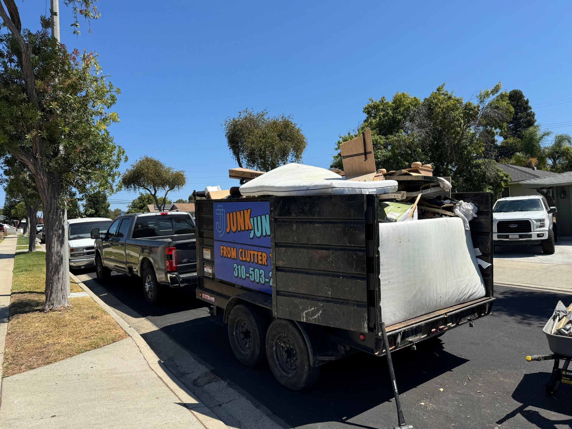 Parked trailer full of junk with colorful sign and white mattress on a sunny street, with cars and trees nearby.