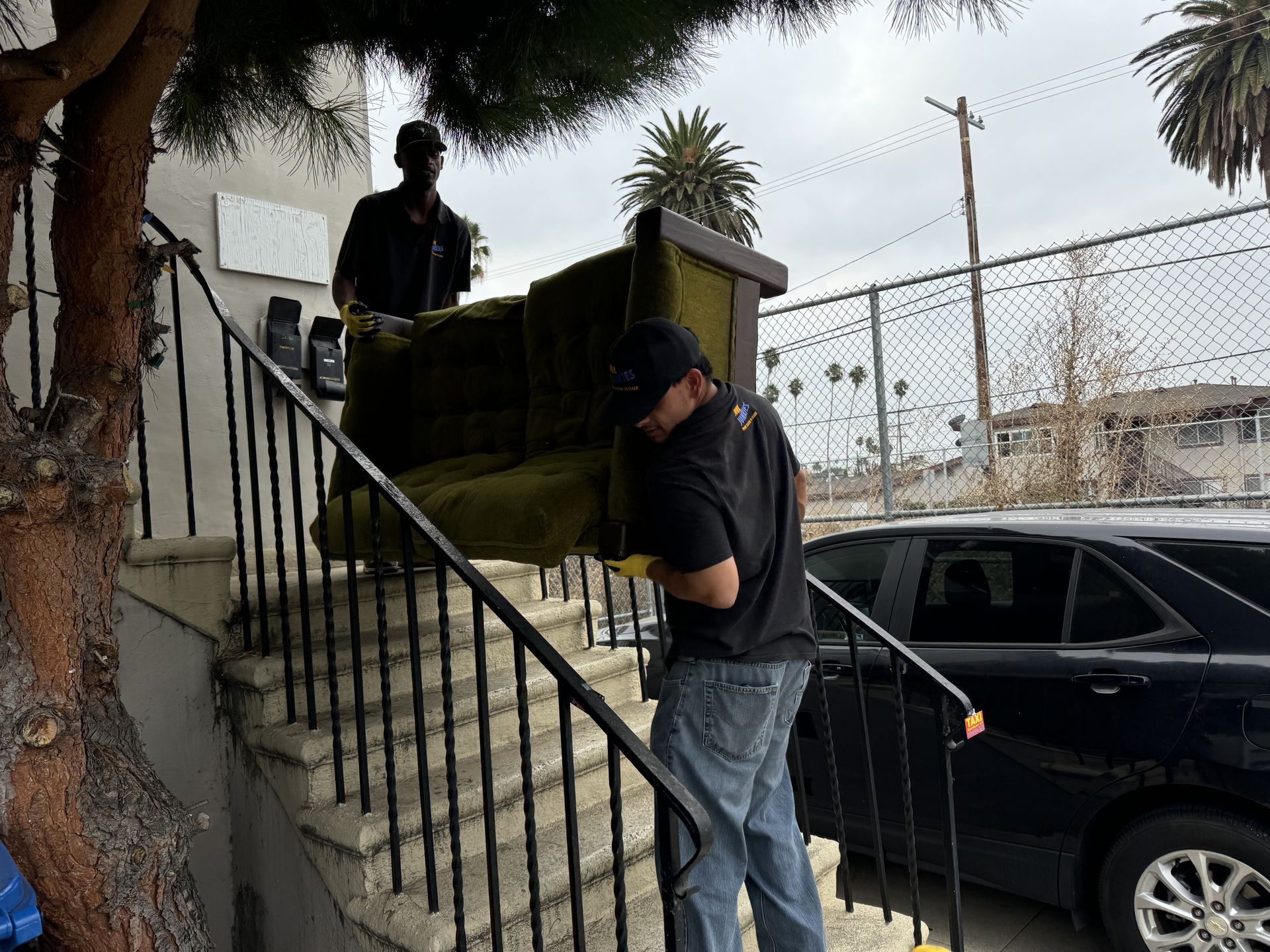 Two people on outdoor stairs holding a couch beside a black car, one standing near the railing and one on the lower steps.