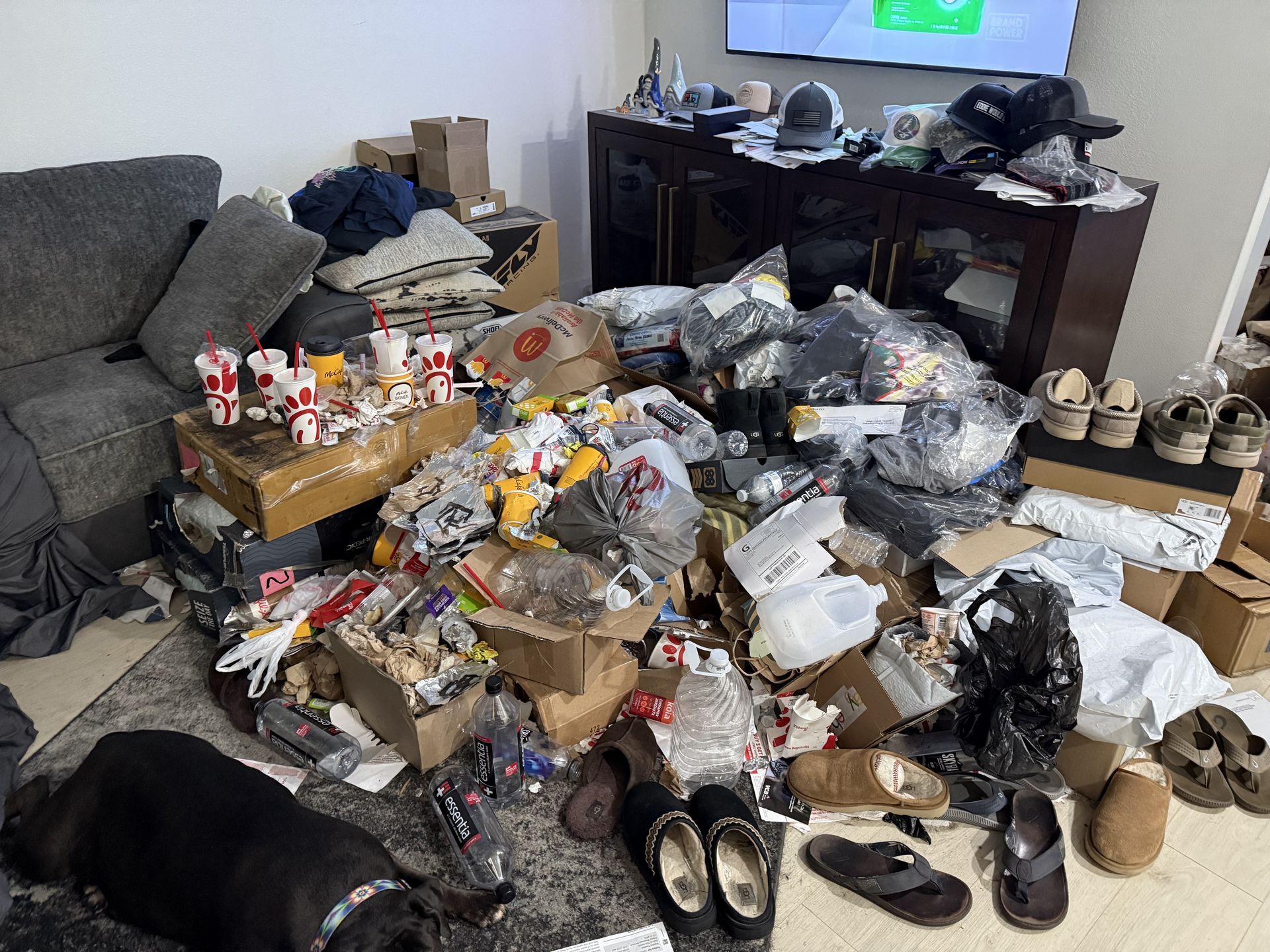 Cluttered living room floor covered with trash bags, boxes, clothes, shoes, and scattered items near a TV.
