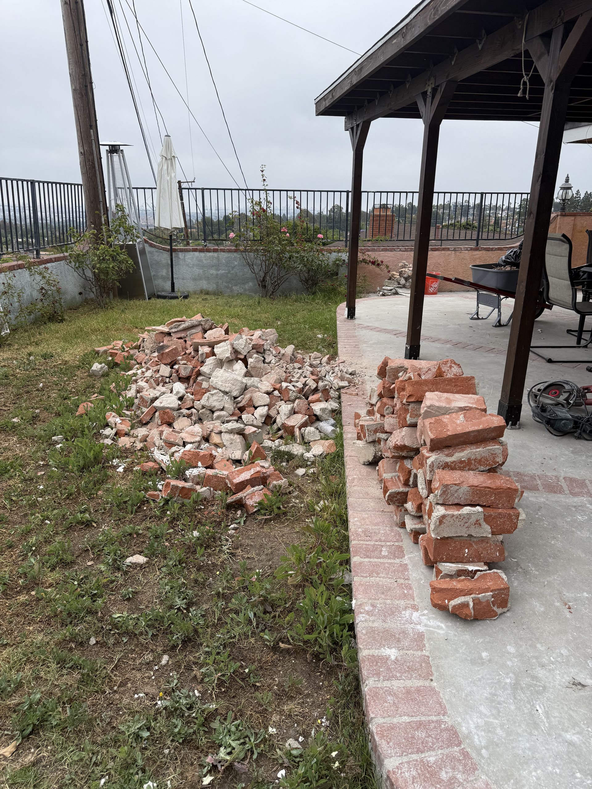 Bricks stacked beside a patio, with a pile of rubble and a grassy yard near a fenced outdoor area.